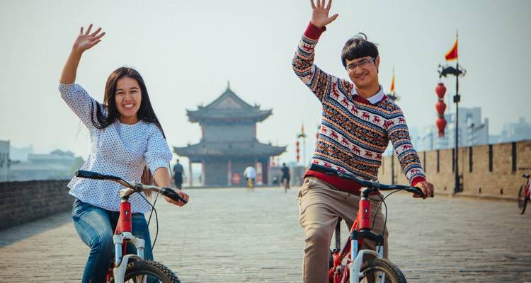 Deux personnes à vélo qui font signe de la main avec une architecture chinoise traditionnelle en arrière-plan.