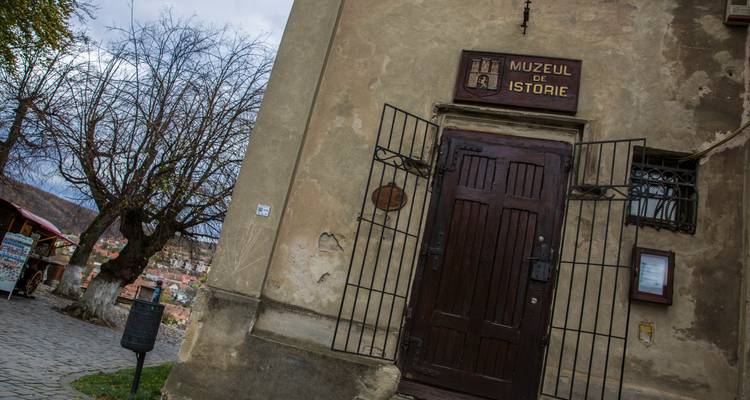 Entrée d'un bâtiment en pierre ancienne avec une porte en bois et un panneau indiquant « Muzeul de Istorie ».