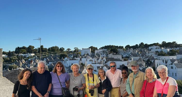 Grupo de turismo sonriente posado en una terraza en la azotea con los tejados de piedra cónicos de los trulli de Alberobello extendidos detrás de ellos bajo un cielo azul despejado.