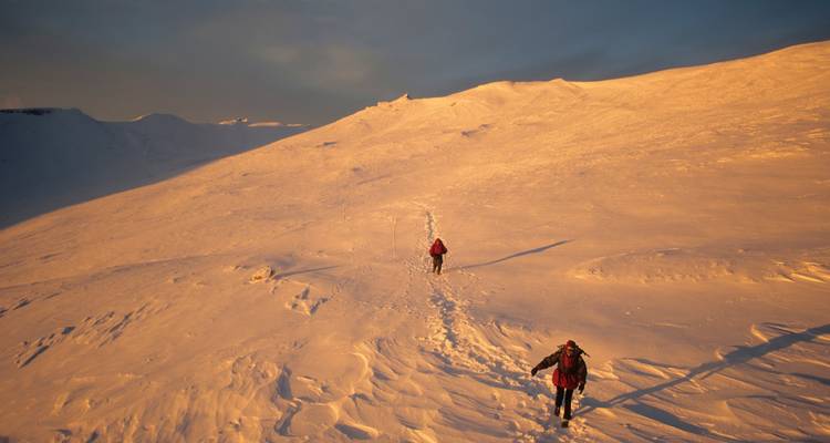 Twee wandelaars wandelen in winterlicht bij zonsondergang op een besneeuwde helling.