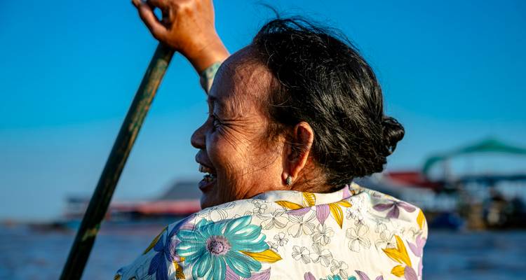Close-up portret van een lachende vrouw die een boot peddelt, felgekleurde bloemenblouse tegen een levendige blauwe lucht.