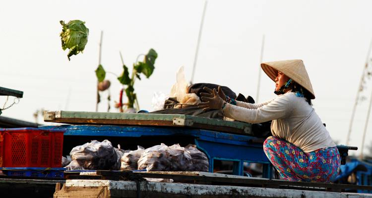 Vrouw op een marktboot gooit groenten terwijl ze gehurkt op een blauw houten dek zit, kegelvormige hoed vangt het licht.