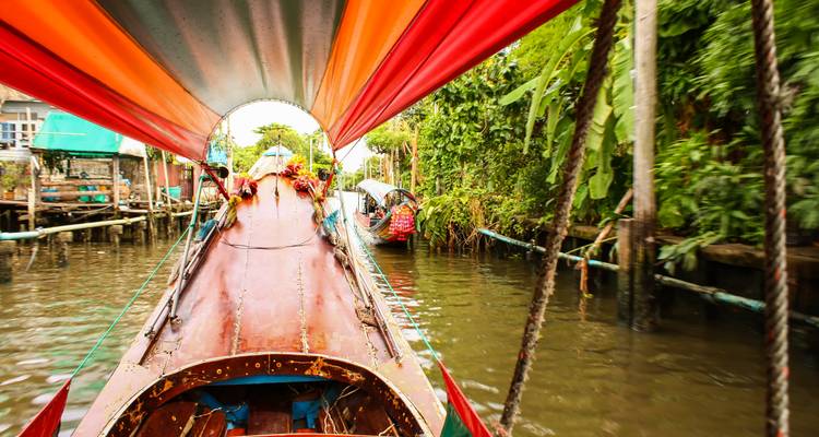 Levendig uitzicht op longtail-boten in een smal kanaal omzoomd met weelderig groen en huizen op stelten.