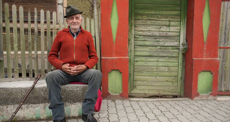 An elderly man sitting on a bench in front of a colorful house.