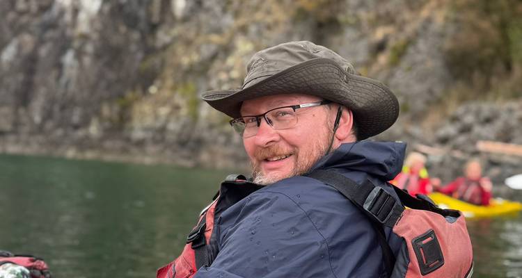 Lächelnder Mann in Regenkleidung und Hut, der in einem Kajak sitzt, mit felsiger Küstenlinie im Hintergrund.