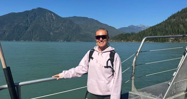 Junge Frau auf dem Bootsdeck mit Rucksack, Berg und grüne Küstenlinie im Hintergrund unter klarem Himmel.