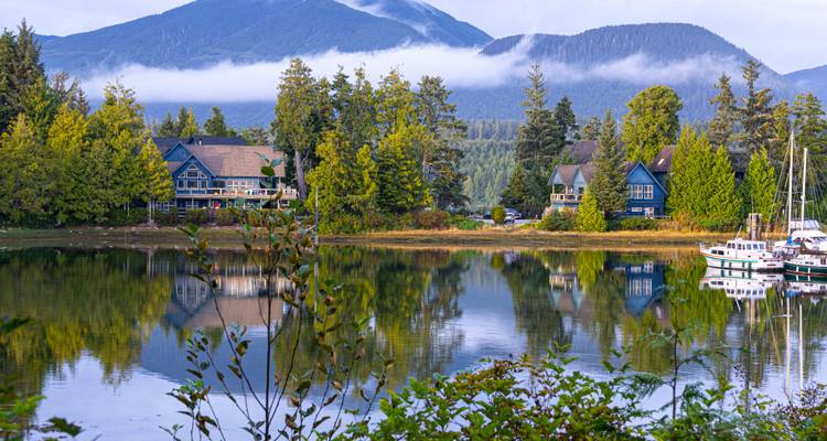 Stille Bucht mit spiegelnden bewaldeten Bergen und rustikalen Lodges bei Tagesanbruch mit leichtem Nebel.