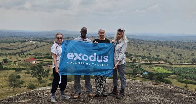 Cuatro viajeros posan en la cima rocosa de una colina sosteniendo un banner de Exodus Adventure Travels con amplias llanuras de fondo.