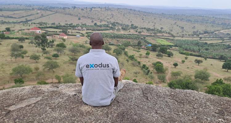 Un hombre con una camiseta de marca se sienta en una roca prominente mirando hacia el amplio paisaje rural de Uganda.