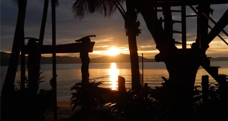 Le coucher de soleil doré se reflète sur la baie tropicale calme, encadré par les silhouettes des arbres et des structures rustiques.