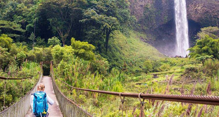 Mochilero cruzando un puente colgante hacia una cascada alta de la selva tropical rodeada de exuberante vegetación.