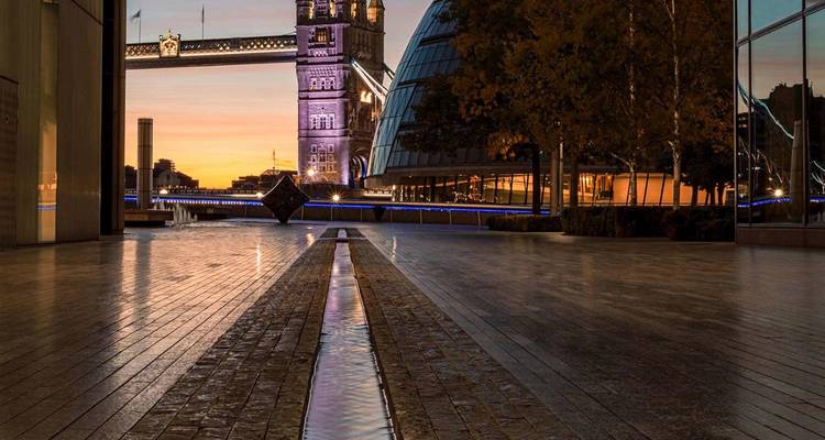 Tower Bridge and modern City Hall glow at dusk beside a wet tiled promenade reflecting the purple-orange sky