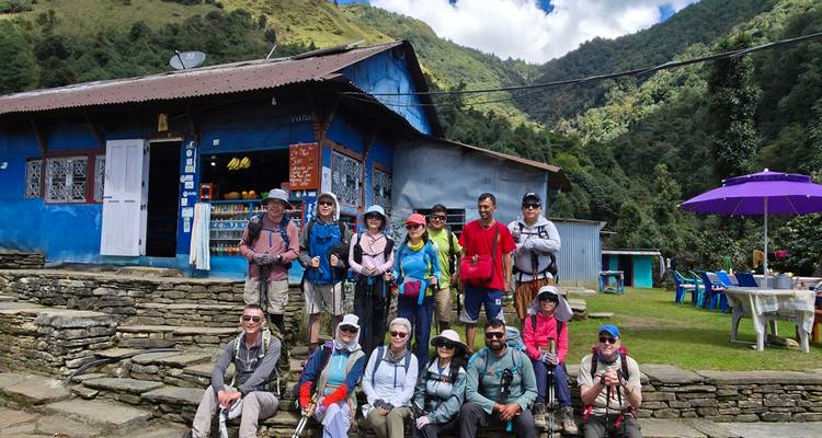 Un groupe de trekking pose avec des sacs à dos devant une maison de thé bleue dans les collines verdoyantes du Népal.