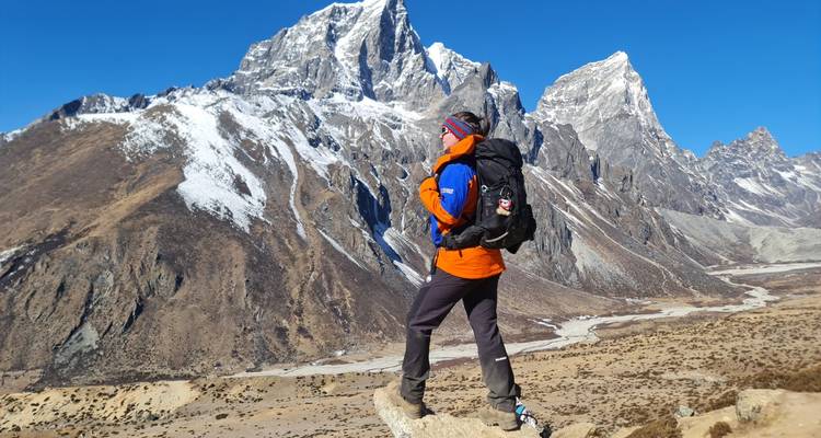 Wandelaar in oranje jas staand op een rotsachtige uitstekende rots met uitzicht op dramatische met sneeuw gestreepte Himalaya-pieken.