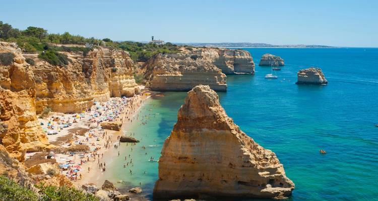 Des falaises de calcaire doré et des pieds de rocher émergeant au-dessus d'une crique turquoise remplie de baigneurs le long du littoral de l'Algarve au Portugal.