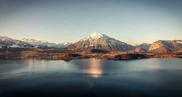 Schneebedeckter Berg, der sich über einen ruhigen See mit kleinen Städten entlang des Ufers bei Morgendämmerung erhebt.