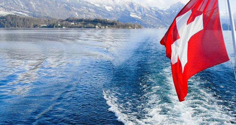 Una bandera suiza ondea detrás de un barco que deja una estela espumosa a través de un profundo lago alpino con montañas nevadas.