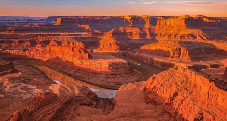 Un vasto paisaje de cañón rojo se curva alrededor de un meandro del Río Colorado bajo un cielo de atardecer resplandeciente en Utah.