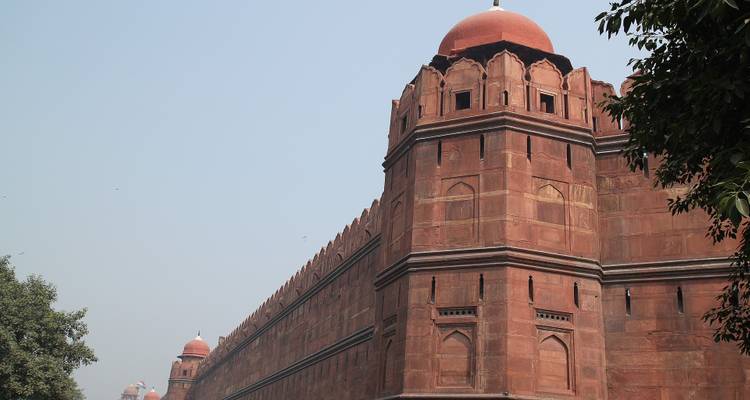 Lange rote Sandsteinmauer der Festung mit Bastionen und gewölbten Türmen, eingerahmt von Bäumen unter einem dunsigen Himmel