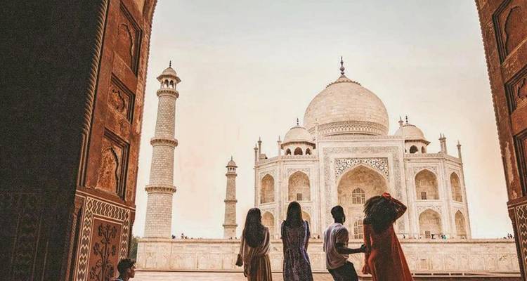 Les silhouettes de cinq visiteurs encadrées par une arche ornementale regardent vers l'emblématique Taj Mahal en marbre blanc à la lumière chaude de l'aube.