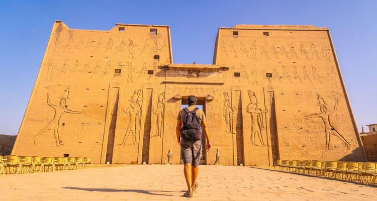Un viajero solitario con una mochila camina hacia la imponente fachada de arenisca cubierta de antiguos relieves egipcios del Templo de Horus.