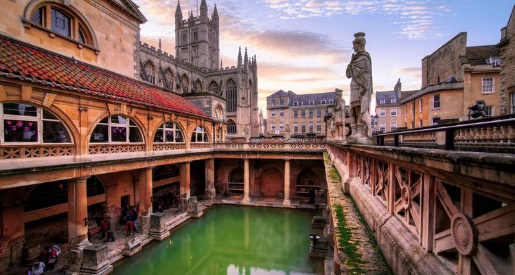 Vista del complejo histórico de los Baños Romanos con la piscina termal verde en primer plano y la Abadía de Bath elevándose detrás bajo un cielo vespertino colorido.
