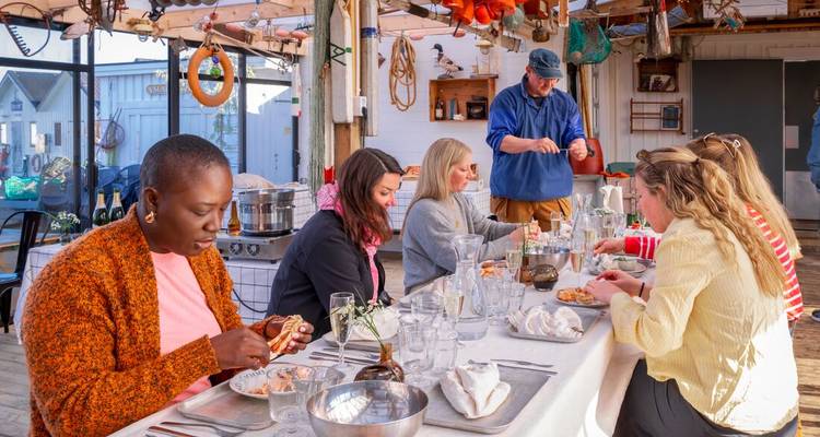 Kleine Gruppe genießt gemeinsam eine Meeresfrüchte-Mahlzeit an einem langen Tisch in einem rustikalen Hafenschuppen.