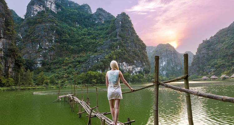 Une personne marchant sur un pont en bambou au-dessus d'un lac serein entouré de montagnes calcaires au coucher du soleil.