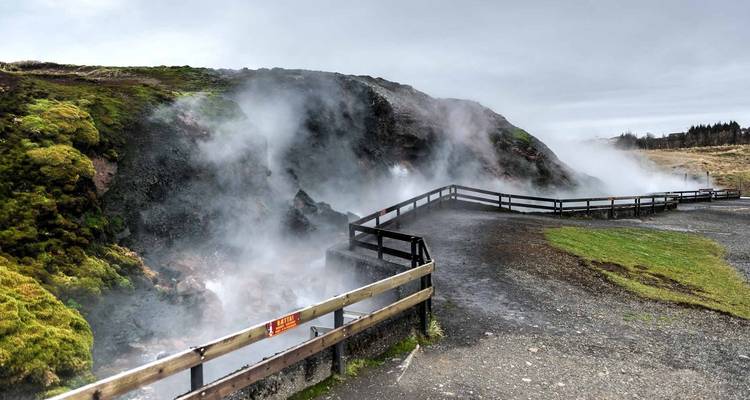 Dampfendes Geothermalgebiet mit einem Holzpfad und Hügeln im Hintergrund.