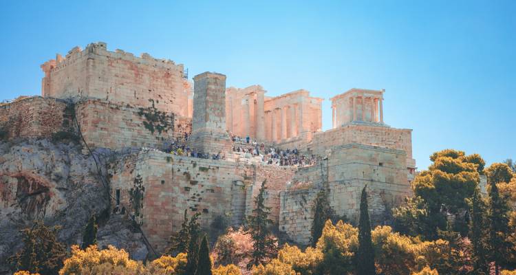 Vista diurna de las murallas de la Acrópolis y multitudes de visitantes vistas desde abajo contra un cielo turquesa brillante.