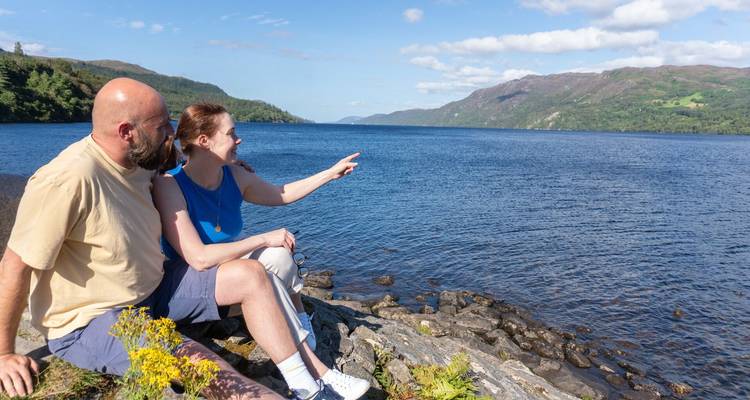 Pareja sentada junto a un lago pintoresco con colinas al fondo.