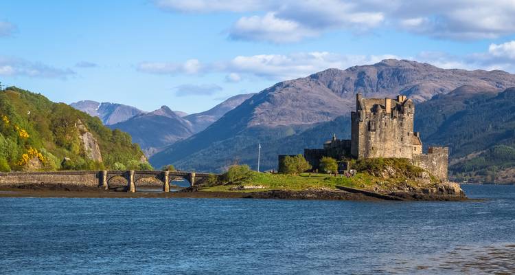 Castillo en una pequeña isla con un puente de piedra y montañas al fondo.