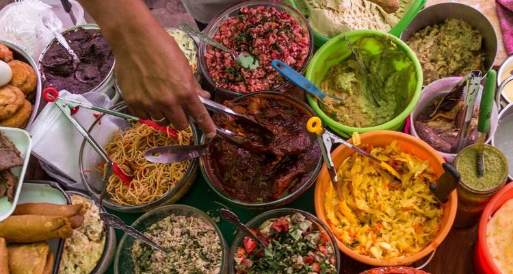 Overhead view of colorful street–food dishes with a vendor’s hand serving from a variety of bowls and pots.