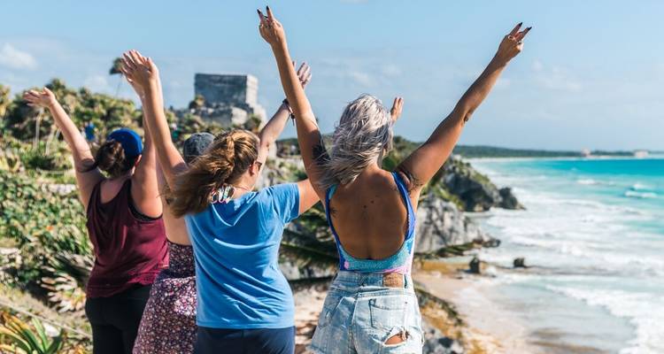 Group of friends with raised arms celebrates on a cliffside near turquoise Caribbean waters and a Mayan ruin tower.