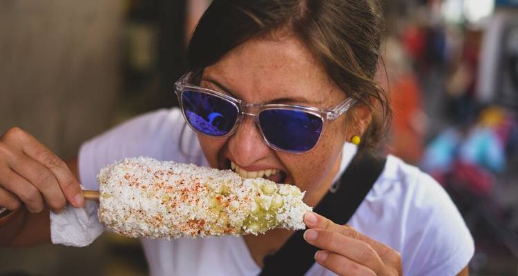 Traveler enthusiastically eating a chili-covered street corn (elote) while wearing reflective sunglasses