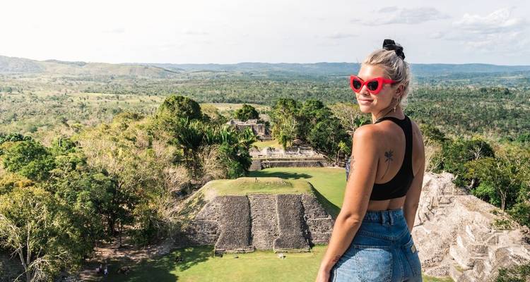 Woman in red sunglasses stands atop ancient stone ruins overlooking jungle and distant temples.