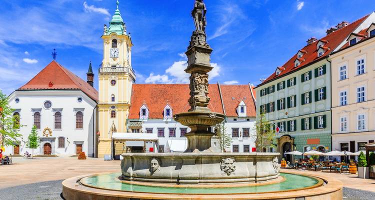 Historischer Brunnen mit Skulpturen steht auf einem hellen gepflasterten Platz in Bratislava, der von pastellfarbenen Gebäuden und einem Glockenturm umgeben ist.