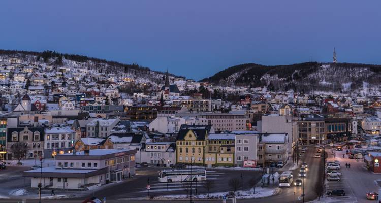 Schneebedeckte skandinavische Stadt zur blauen Stunde mit leuchtenden Lichtern und Hügeln im Hintergrund