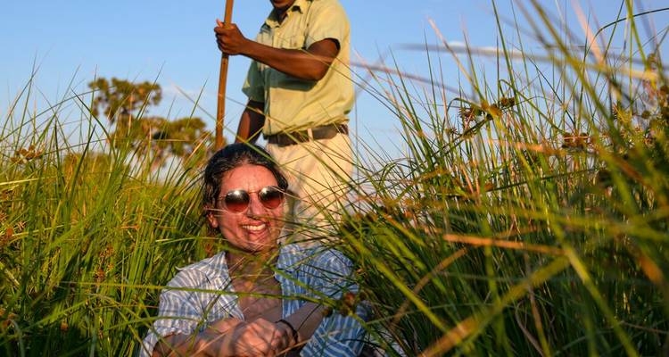 Viajera sonriendo entre cañas altas mientras un barquero navega una canoa tradicional detrás de ella al atardecer.