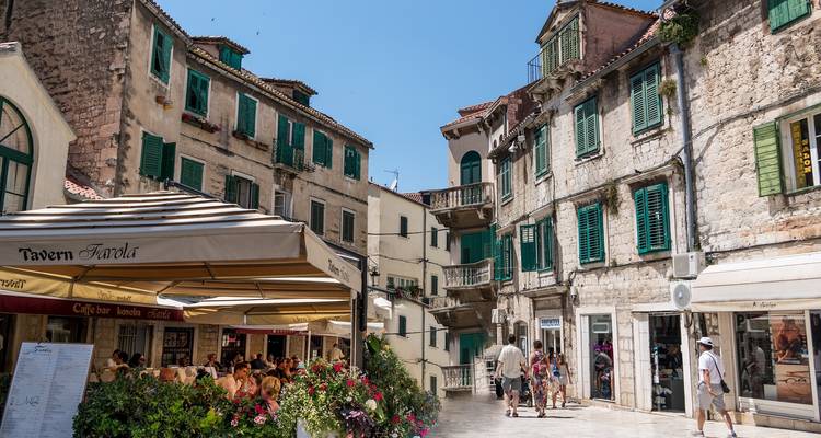 Ruelle pavée animée dans une vieille ville méditerranéenne avec des terrasses de café et des bâtiments aux volets verts