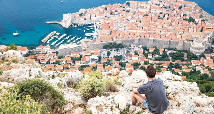 Un voyageur se repose sur un promontoire rocheux au-dessus des toits de terre cuite et des murs médiévaux d'une ville côtière