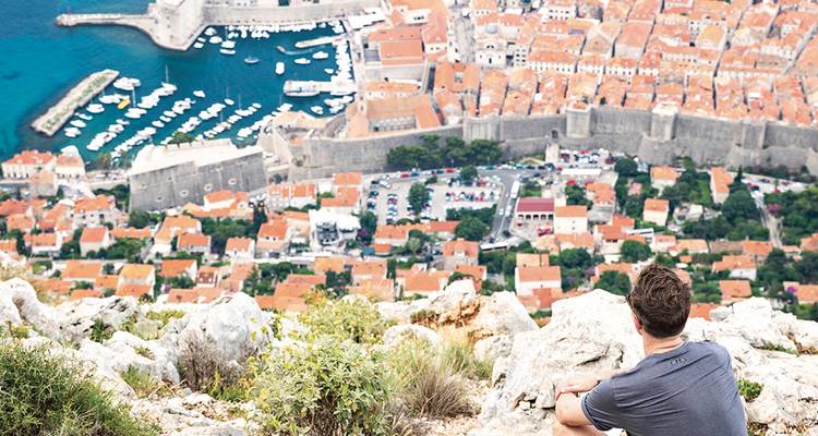 Un voyageur est assis au sommet d'une colline rocheuse surplombant la vieille ville fortifiée de Dubrovnik et le port adriatique.