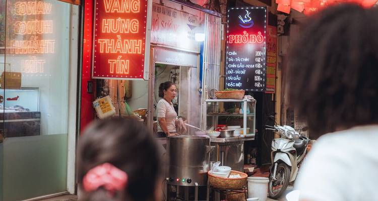 Étal de rue nocturne avec enseignes au néon où un vendeur prépare un pho fumant