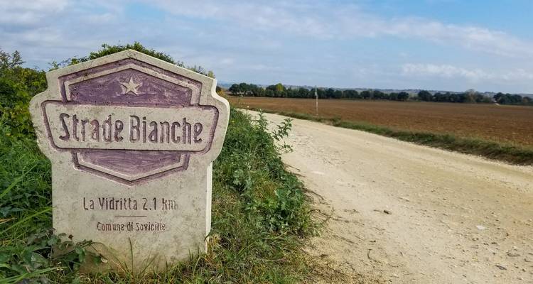 Versleten wegwijzer met 'Strade Bianche' erop langs een grindpad door het platteland.