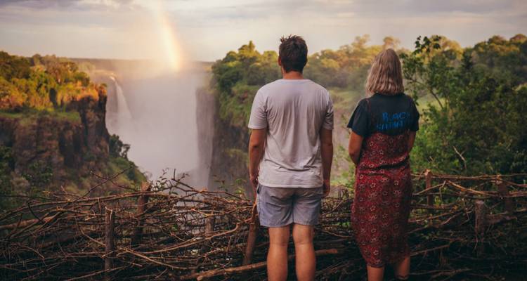Un couple debout observant les chutes Victoria avec un arc-en-ciel.