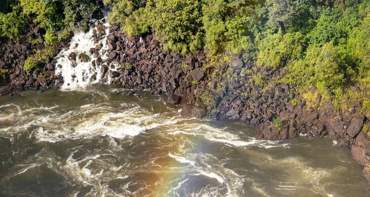 Vista aérea de rápidos turbulentos de río debajo de una cascada con un arcoíris tenue y exuberante selva circundante.