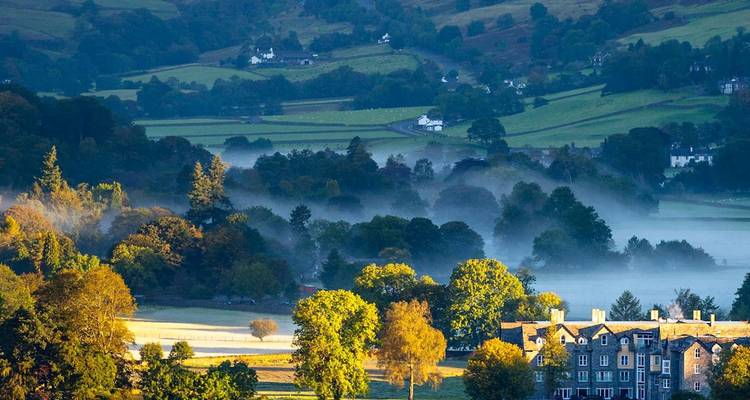 La niebla se alza sobre un tranquilo valle verde salpicado de árboles, casas de campo y un lago reflectante, rodeado de suaves colinas ondulantes