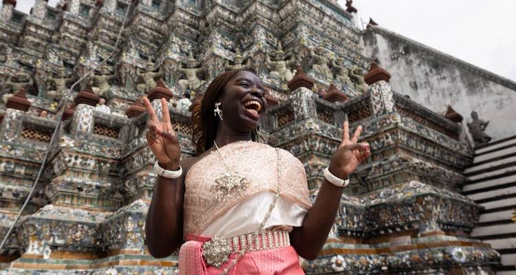 Mujer sonriente en vestimenta tradicional tailandesa posando con signos de victoria frente a la arquitectura ornamentada de un templo