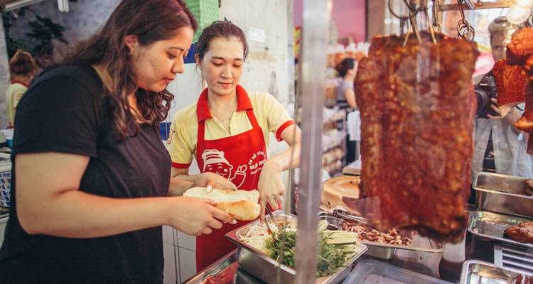 Local vendor prepares a baguette sandwich for a traveller at a busy street food stall.