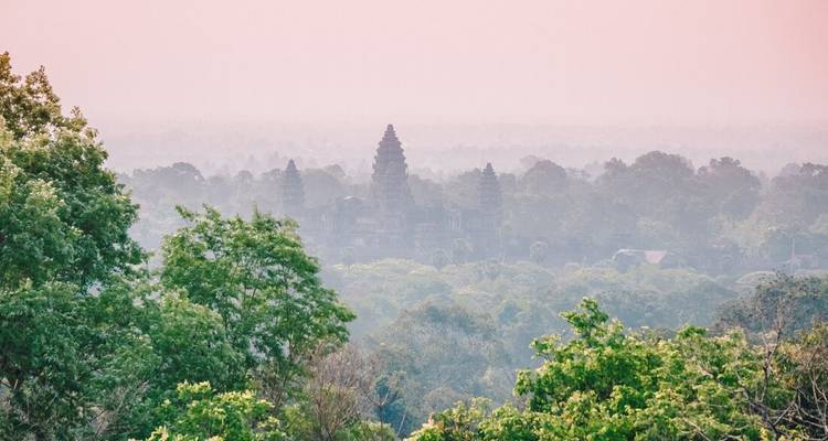 Misty pink sunrise view over the treetops revealing distant towers of Angkor Wat.